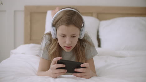 Kleines-Mädchen-Spielt-Online-Spiel-Auf-Dem-Smartphone-Und-Tippt-Auf-Den-Touchscreen,-Liegt-Im-Schlafzimmer-Und-Hört-Ton-über-Kopfhörer
